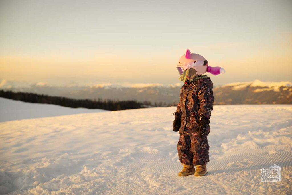 Kind steht in Winter mit Schnee am Berg, bekleidet mit Skianzug, Helm. Am Helm sind rosa Plüschohren und ein rosa Fuchsschwanz aufgeklebt. Die Abendsonne färbt die Szene in ein warmes Licht.